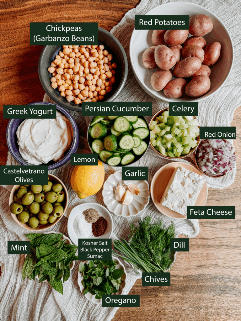 Ingredients for tzatziki potato salad including red potatoes, cucumbers, chickpeas, feta, olives, and fresh herbs.