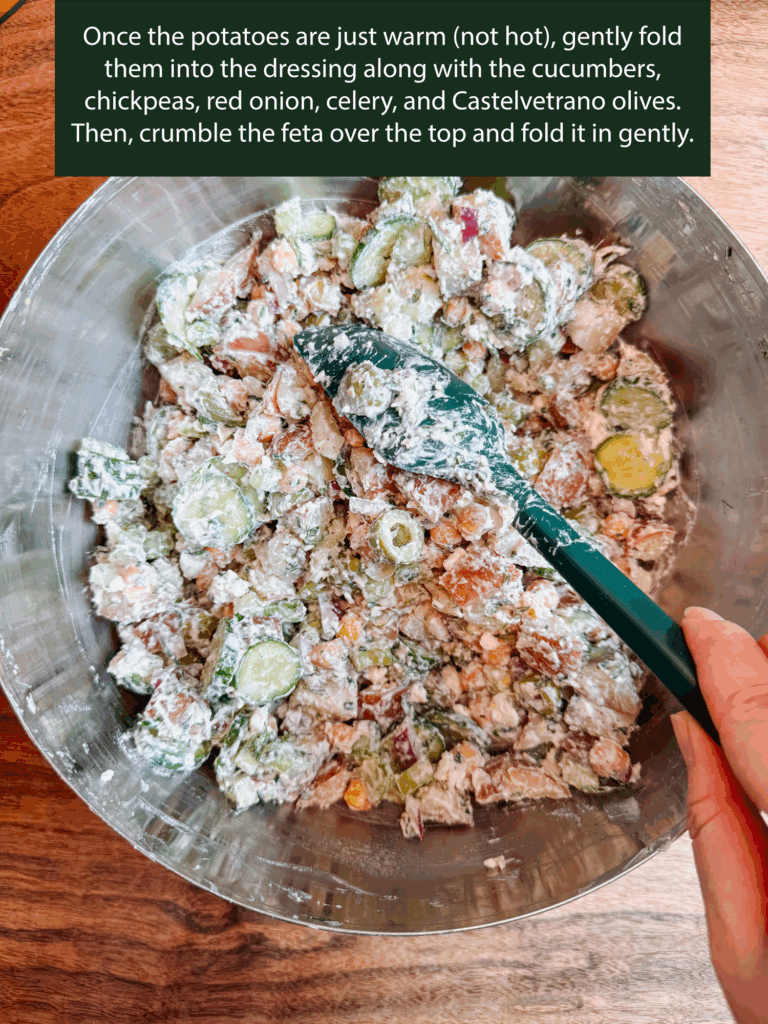 Mixing tzatziki potato salad with cucumbers, chickpeas, red onion, celery, and olives in a metal bowl with a green spatula.