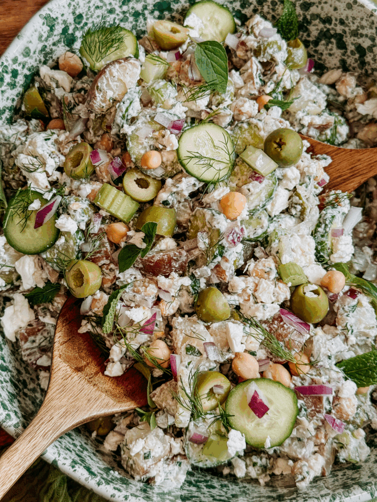 Close-up of tzatziki potato salad with cucumbers, olives, chickpeas, and feta in a serving bowl with wooden spoons.