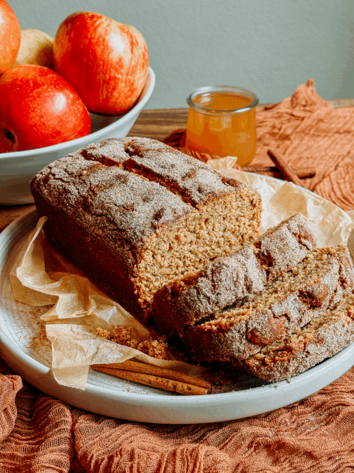 Sliced apple cider donut bread with a crisp cinnamon-sugar crust on a parchment-lined plate with apples and apple cider in the background.