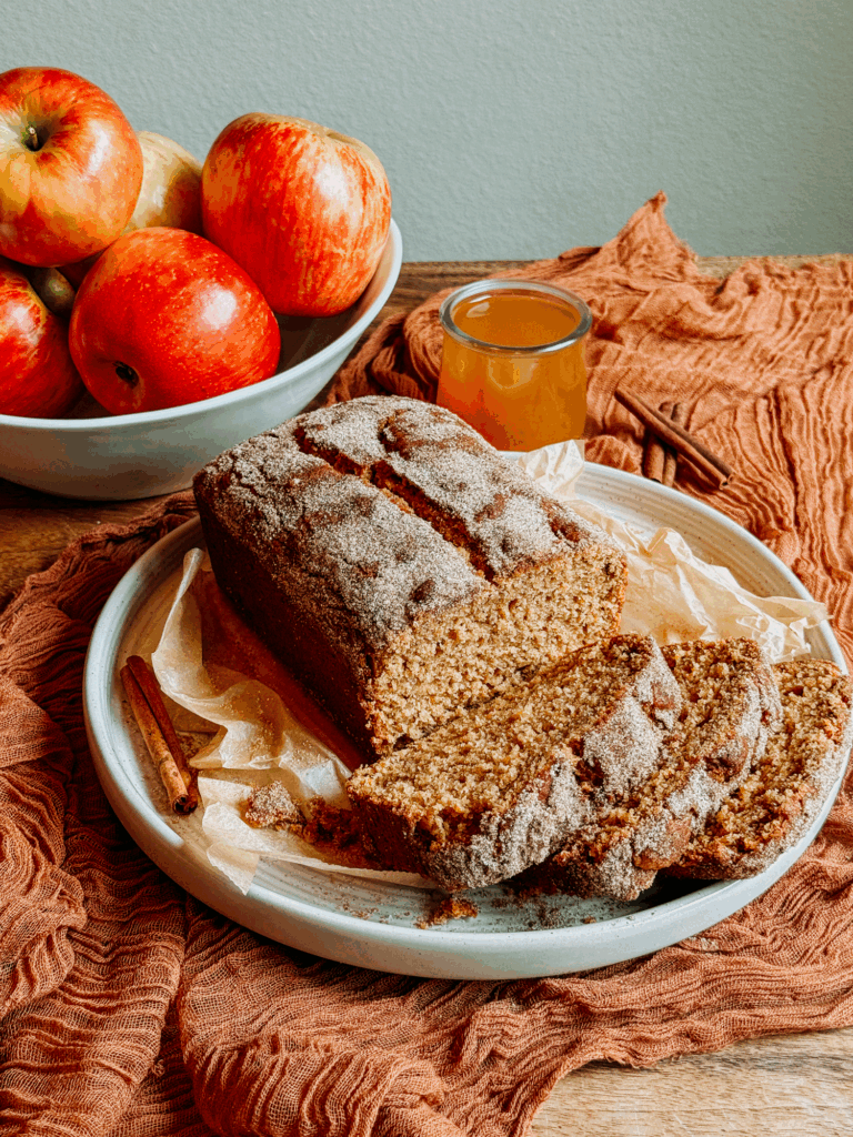 Sliced apple cider donut bread on a plate with fresh apples, cinnamon sticks, and apple cider.