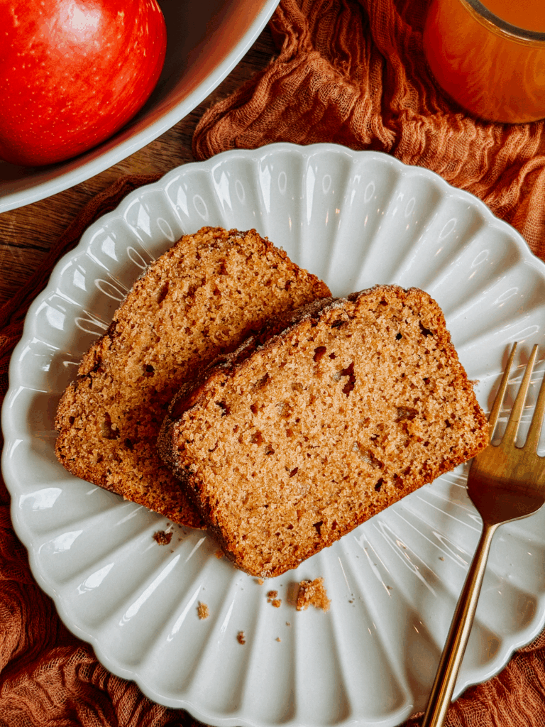 Two slices of apple cider donut bread on a scalloped white plate with a gold fork.