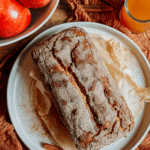 Whole loaf of apple cider donut bread on a white plate with a sugar crust, surrounded by apples, cinnamon sticks, and a small glass of apple cider.