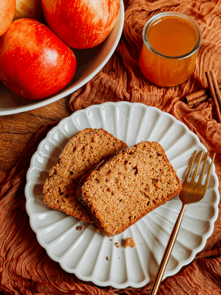 Two slices of apple cider donut bread on a white plate with apples and cider.