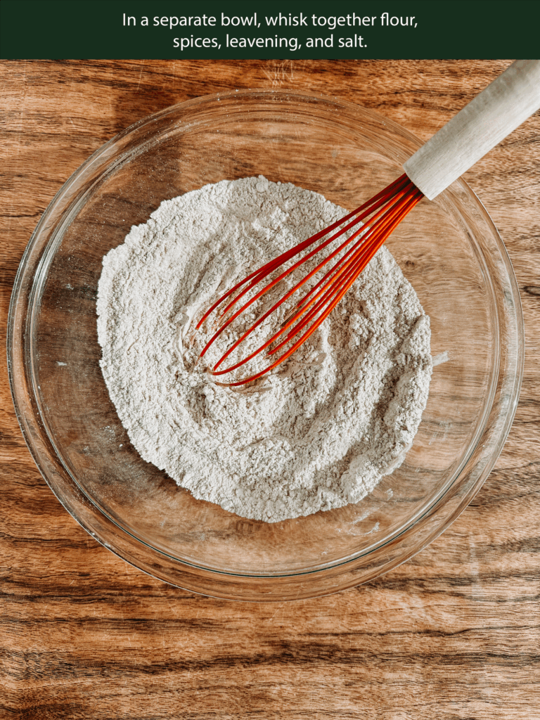 Bowl of dry ingredients with whisk including flour, cinnamon, cardamom, nutmeg, ginger, baking powder, and salt.