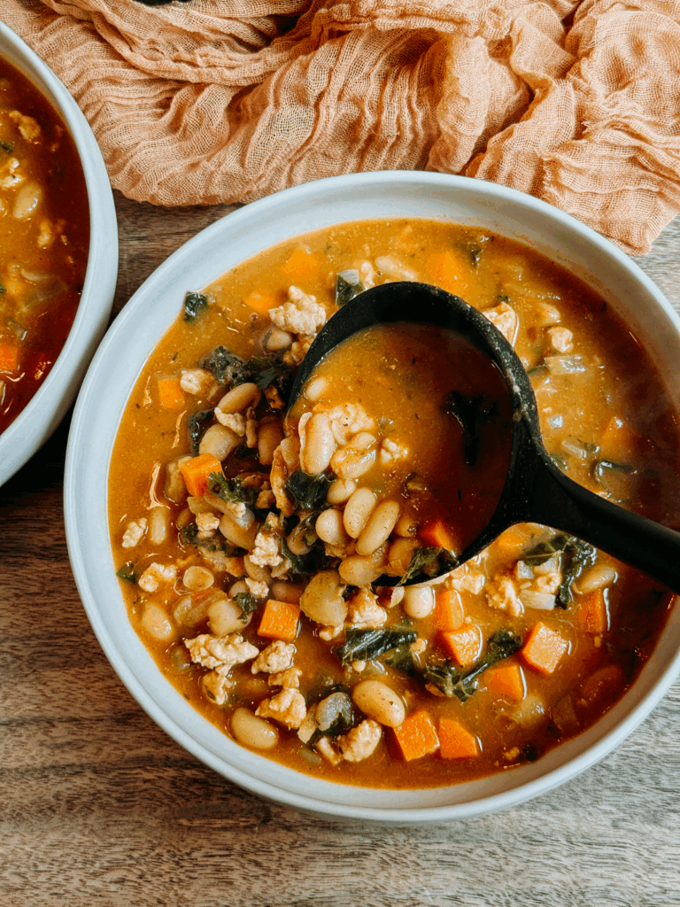 Close-up of a ladle lifting a hearty scoop of Turkey Pumpkin White Bean Chili with cannellini beans, kale, turkey, and carrots.