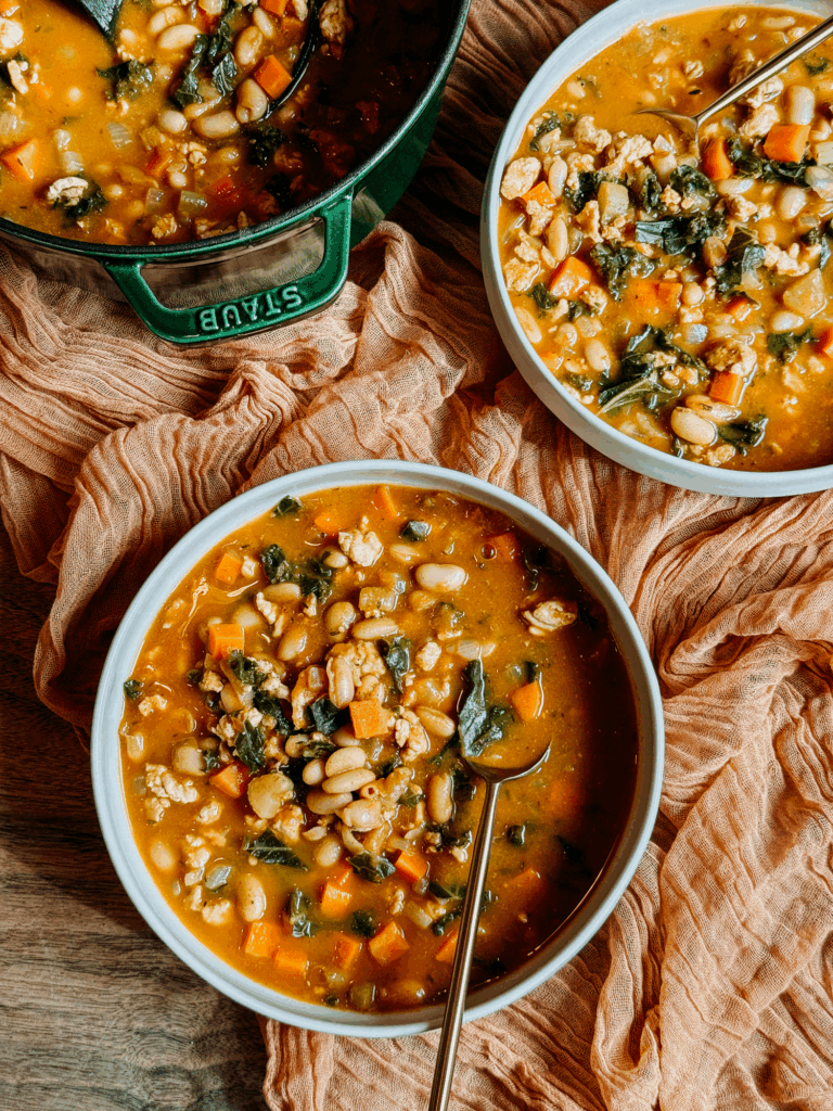 Overhead view of two bowls of Turkey Pumpkin White Bean Chili next to a Dutch oven, showcasing beans, vegetables, and kale in the pumpkin broth.