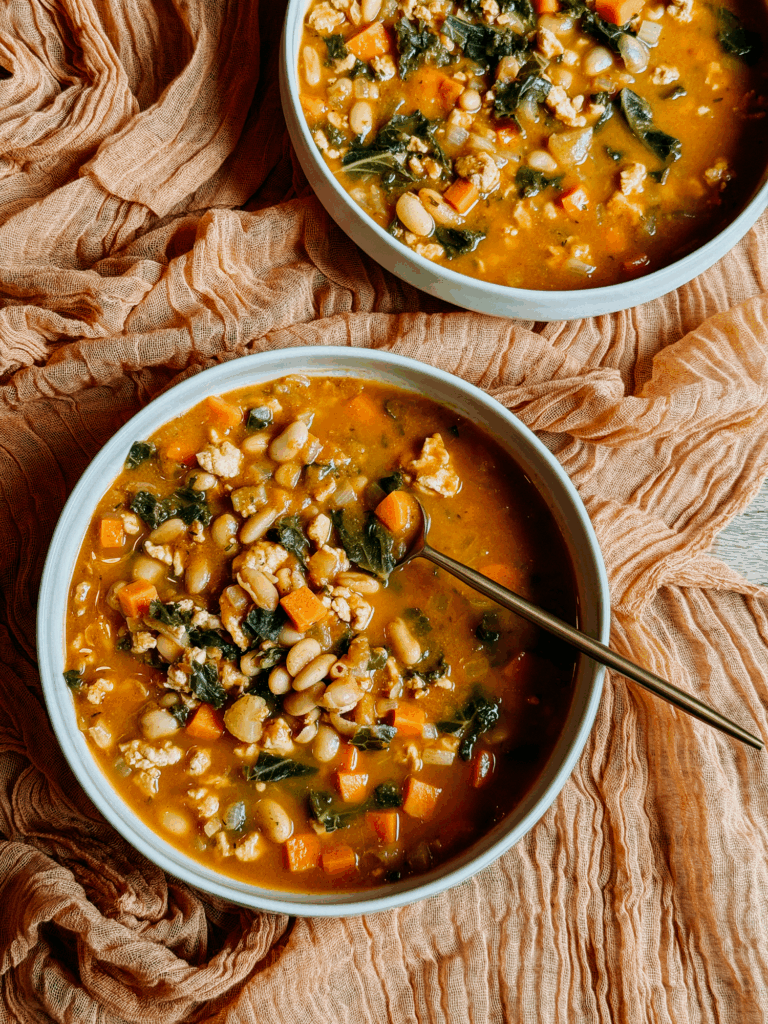 Two bowls of Turkey Pumpkin White Bean Chili arranged on a textured orange cloth, filled with beans, vegetables, kale, and turkey.