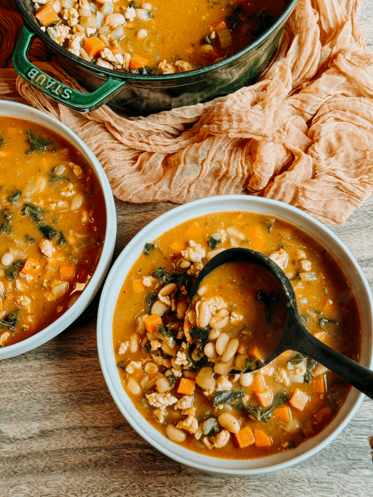 Ladle serving warm Turkey Pumpkin White Bean Chili filled with beans, carrots, kale, and ground turkey from a green Dutch oven into bowls.
