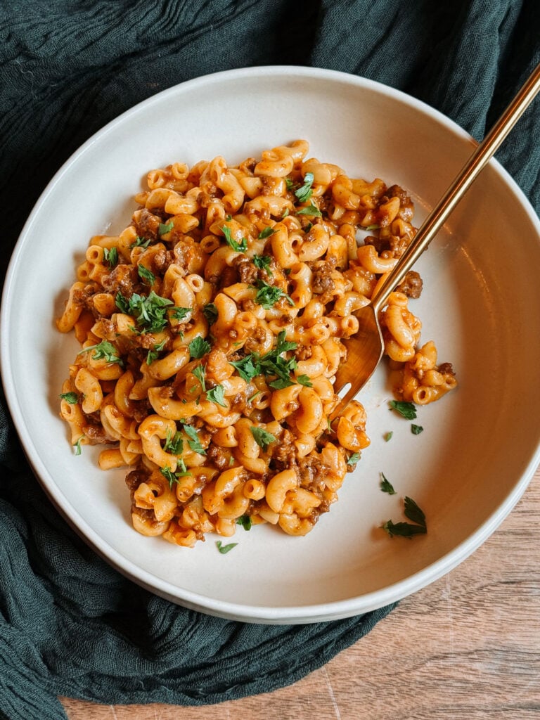 A full bowl of homemade cheeseburger macaroni with ground beef, creamy cheddar sauce, and parsley on dark green linen.