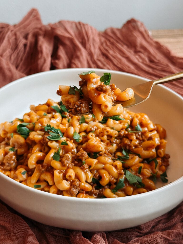 A fork lifting a bite of cheesy beef macaroni above a bowl of homemade Instant Pot Hamburger Helper with parsley.