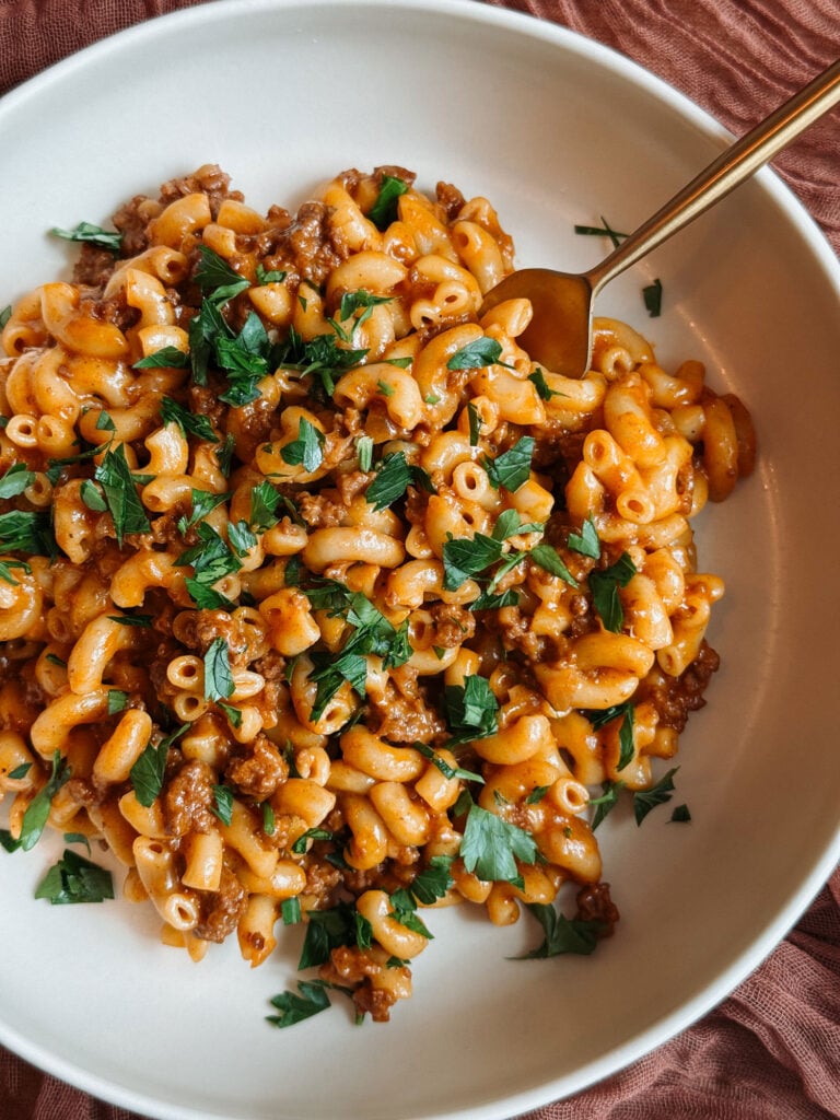 Overhead view of cheeseburger macaroni with ground beef, creamy cheddar sauce, chopped parsley, and a gold fork in a white bowl.